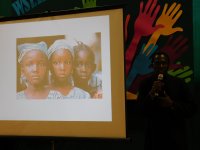 Przemawia ks. Jan z Kamerunu, który za pomocą prezentacji multimedialnej, przedstawia życie w Kamerunie. Na slajdzie wyświetlone jest zdjęcie trzech ciemnoskórych dziewczynek, dwie z nich mają zawiązane chustki na głowie.
