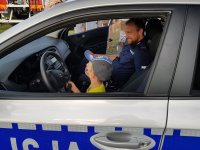 Umundurowany Policjant, siedzi w radiowozie z chłopcem, któremu objaśnia działanie wyposażenia pojazdu.