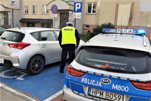 Policjant Wydziały Ruchu Drogowego sprawdza prawidłowość zaparkowanego pojazdu na miejscu wyznaczonym dla osób niepełnosprawnych. Po prawej stronie zaparkowany, oznakowany radiowóz.