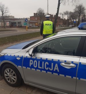 Na pierwszym planie oznakowany radiowóz, tuż za nim stoi umundurowany policjant obserwuje zachowanie uczestników ruchu drogowego.