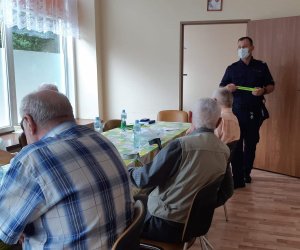 Umundurowany policjant rozmawia z seniorami siedzącymi w sali Dziennego Pobytu Opieki Medycznej w Zambrowie.