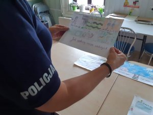 Umundurowana policjantka ocenia prace wykonane na konkurs plastyczny  pod nazwą ,,Bezpieczne wakacje 2020&quot;. W reku trzyma plakat przedstawiający różne formy wypoczynku.