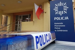 Podświetlony napis POLICJA, na dachu radiowozu. Po lewej stronie zdjęcia logo Komendy Powiatowej Policji w Zambrowie, na niebieskim tle. Na drugim planie wejście do budynku komendy.