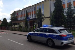 Policjant drogówki w rejonie szkoły podstawowej numer cztery, sprawdza prawidłowość oznakowania przejścia dla pieszych. Na pierwszym planie oznakowany radiowóz policyjny.