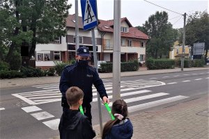 Policjant przekazuje opaski odblaskowe przechodzącym dzieciom. W tle zdjęcia przejście dla pieszych.