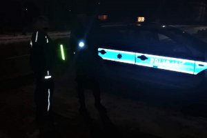 Noc. Policjant przekazuje opaskę odblaskowa przechodniowi, która odbija światło. Po prawej stronie zdjęcia oznakowany radiowóz.