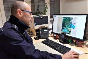 Umundurowany policjant siedzi przed komputerem podczas spotkania online. Na monitorze komputera widoczna ulotka z ostrzeżeniami dla wolontariuszy.