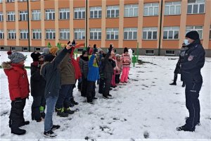 Po prawej stronie zdjęcia policjantka, po lewej uczniowie. W tle budynek szkoły.