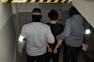 Dwóch policjantów wydziału kryminalnego wraz zatrzymanym mężczyzna, który ma zapięte kajdanki na ręce trzymane z tyłu. w tle zdjęcia krata.
