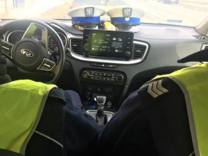 Zdjęcie zrobione w radiowozie policyjnym, w pojeździe dwóch policjantów w żółtych kamizelkach, a na podszybiu dwie czapki policyjne.