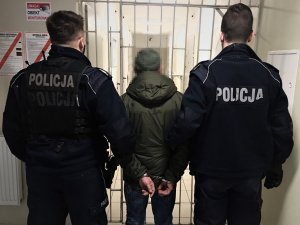 Dwóch funkcjonariuszy policji podczas doprowadzania zatrzymanego, który na zapięte kajdanki na rękach trzymanych z tyłu. W tle kraty.