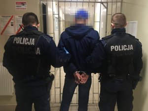 Dwóch umundurowaniach policjantów, a pomiędzy nimi zatrzymany w kajdankach na rekach trzymanych z tyłu podczas doprowadzenia.