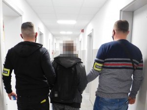 Na środku zatrzymany, a po jego bokach policjanci z wydziału kryminalnego w  odzieży cywilnej z opaskami na przedramionach  z napisem policja, podczas doprowadzenia. W tle długi, wąski korytarz.