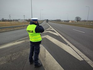 Policjant ruchu drogowego w żółtej kamizelce stojący na drodze tyłem podczas pomiaru prędkości. W tle jezdnia oraz pojazdy.