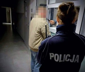 Umundurowana policjantka stojąca tyłem, patrząca na mężczyznę badanego na zawartość alkoholu w wydychanym powietrzu. W tle korytarz.