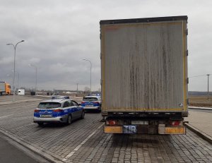 Z prawej strony tył stojącego na parkingu tira. Z lewej strony stoją dwa oznakowane radiowozy policyjne. W tle parking, latarnie.