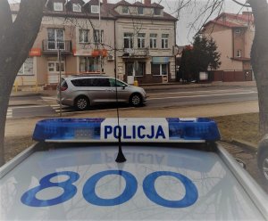 Dach radiowozu z światłami z napisem Policja, w tle ulica i jadący nią samochód osobowy koloru srebrnego oraz zabudowania.