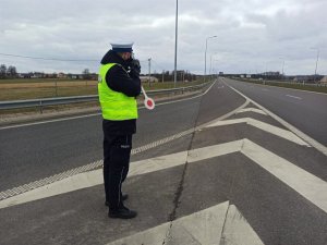 Umundurowany policjant stojący bokiem na drodze ekspresowej podczas pomiaru prędkości. W tle jezdnia, a nią jadące samochody oraz zabudowania i pola na poboczu.