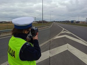 Umundurowana policjanta stojący bokiem na drodze ekspresowej podczas pomiaru prędkości. W tle jezdnia, a nią jadące samochody oraz zabudowania i pola na poboczu.