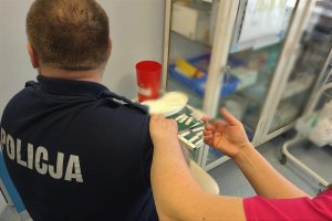 Z prawej strony pielęgniarka podczas szczepienia policjanta, który jest odwrócony plecami. W tle szafka.