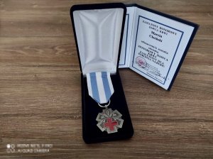 Na biurku leży w pudełku medal w kształcie gwiazdy, a środku czerwony krzyż i kwiat lili oraz biało- niebieska szarfa. Z tyłu postawiona książeczka z tekstem &quot;ZASŁUŻONY HONOROWY DAWCA KRWI Marcin Choiński odznaczony/a został/a odznaką HONOROWY DAWCA KRWI ZASŁUŻONY DLA ZDROWIA NARODU&quot;. Na dole pieczątka o tekście &quot;Z upoważnienia MINISTRA ZDROWIA PODSEKRETARZ STANU Sławomir Gadowski &quot; oraz własnoręczny podpis. W prawym dolnym rogu &quot;Minister Zdrowia&quot;.