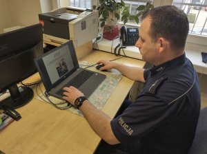 Policjant podczas spotkania on line. Siedzi przed biurkiem, patrzy na ekran laptopa, na którym wyświetlona jest prezentacja, a na niej tytuł &quot;STOP PRZEMOCY!!!&quot; oraz zdjęcie. W tle sprzęt biurowy.