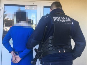 Z prawej strony umundurowany policjant trzymający doprowadzana kobietę za przedramię. Kobieta ma założone kajdanki na ręce trzymane z tyłu. w tle szklane drzwi.