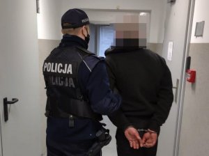 Po prawej stronie zatrzymany z rękoma trzymanymi z tyłu i zapiętymi kajdankami, którego doprowadza umundurowany policjant trzymający do za przedramię.