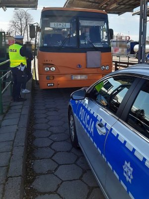 Stojący na dworcu pks autobus podczas kontroli policjanta, który stoi przed jego drzwiami. Przed autobusem zaparkowany radiowóz policyjny.