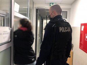Z prawej strony umundurowany policjant, a obok niego kobieta podczas badania na zawartość alkoholu w organizmie. W tle korytarz oraz szklane okna oddzielające dyżurnego.