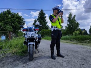 Po prawej stronie policjant podczas pomiaru prędkości, obok niego motor policyjny.