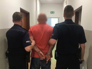 Po bokach umundurowani policjanci podczas doprowadzenia zatrzymanego, którego trzymają za przedramiona pomiędzy nimi. Zatrzymany ma założone kajdanki na ręce trzymane z tyłu. W tle korytarz i drzwi.