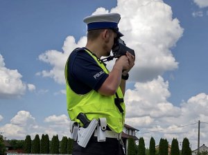 Policjant w kamizelce odblaskowej podczas pomiaru prędkości. W tle krzewy oraz niebo.