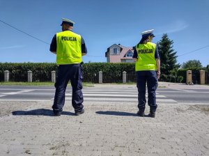 Policjanci w kamizelkach odblaskowych przed przejściem dla pieszych. Zdjęcie robione  z tyłu.