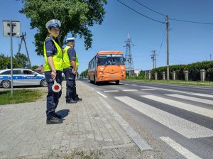 Policjanci w kamizelkach odblaskowych przed przejściem dla pieszych. W tle radiowóz.