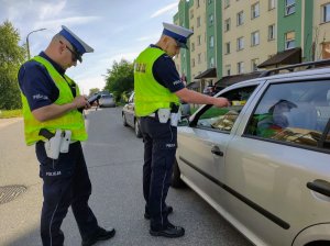 Policjant bajający stan trzeźwości kierującego siedzącego w srebrnym pojeździe. obok policjanty wykon ujący czynności służbowe na urządzeniu mobilnym.