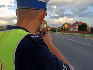 Policjant podczas pomiaru prędkości , w tle droga oraz zabudowania.