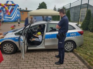 Policjant z dzieckiem w radiowozie pokazujący mu sprzęt służbowy. Przed radiowozem stoi drugi policjant.