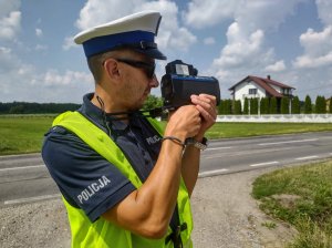 Umundurowany policjant wydziału ruchu drogowego podczas pomiaru prędkości. W tle jezdnia.