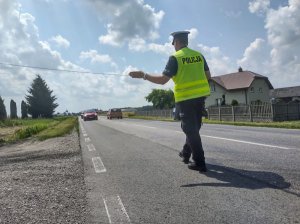 Umundurowany policjant wydziału ruchu drogowego wskazujący jadącemu samochodowi miejsce zatrzymania. W tle jezdnia.