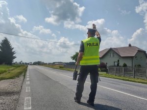 Umundurowany policjant wydziału ruchu drogowego wskazujący jadącemu samochodowi znak zatrzymania poprze uniesienie ręki w górę. W tle jezdnia.