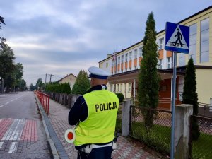 Policjant stojący na chodniku przed przejściem dla pieszych przy szkole.