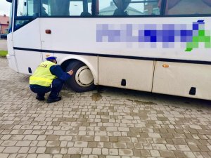 Umundurowany policjant podczas kontroli autobusu, sprawdza ogumienie.