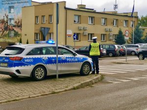 Policjant stojący prze radiowozie obserwujący ruch na przejściu dla pieszych. w tle zabudowania.