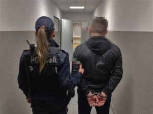 Umundurowania policjantka podczas doprowadzenia zatrzymanego, który ma założone kajdanki na ręce trzymane z tyłu.