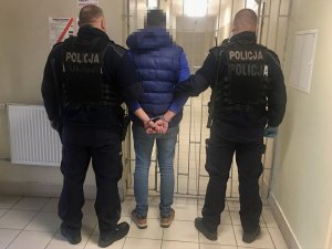 Umundurowani policjanci podczas doprowadzenia zatrzymanego, który ma założone kajdanki na ręce trzymane  z tyłu.