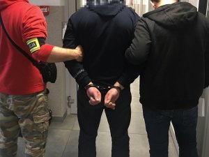 Policjanvi wydziału kryminalnego podczas doprowadzenia zatrzymanego, który ma założone kajdanki na ręce trzymane z tyłu.