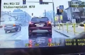 Widok z kamery pojazdu policji. Na ekranie cyfry w górnej i dolnej części obrazu. Skrzyżowanie z sygnalizacją świetlną, srebrny samochód po lewej stronie wjeżdża 
za sygnalizator wyświetlający czerwone światło i skręca w lewo.