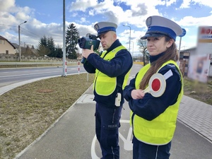 Policjant podczas pomiaru prędkości na ulicy, obok stojąca policjanta z tarczą do zatrzymywania pojazdów.