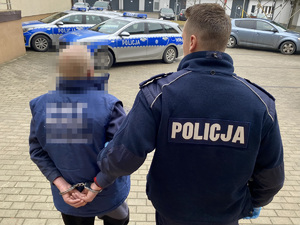 Policjant podczas doprowadzenia zatrzymanego, który ma złożone kajdanki na ręce trzymane z tyłu.